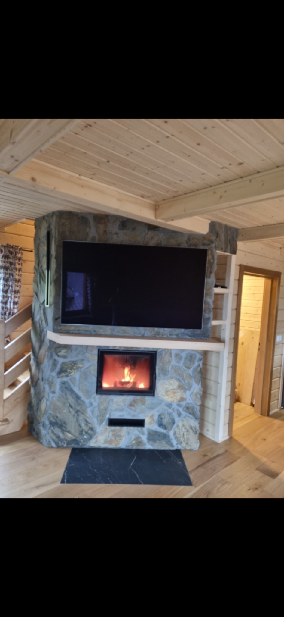 Salon w stylu rustykalnym z kamiennym kominkiem, w którym umieszczony jest telewizor. Drewniane wykończenie sufitu i ścian, podłoga z paneli. W kominku płonie ogień.