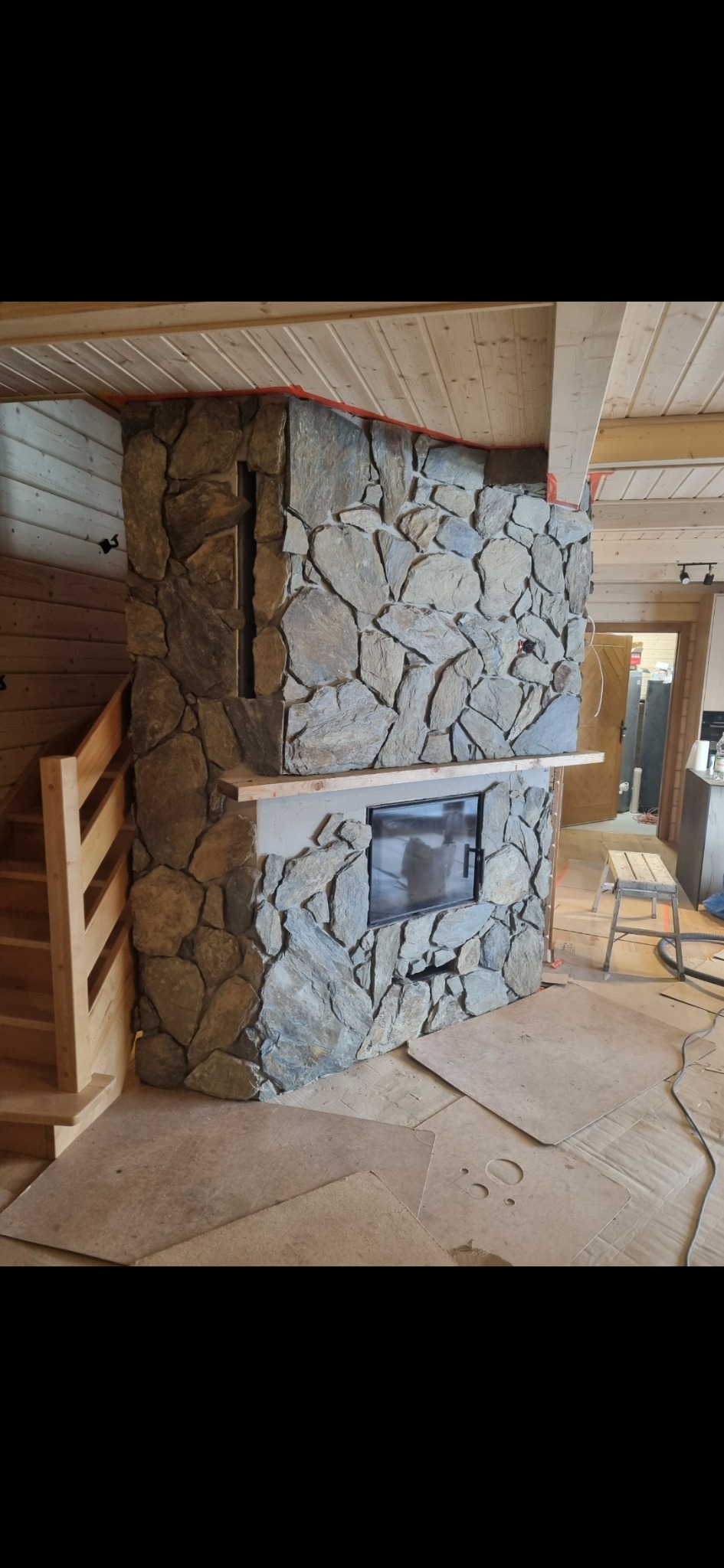 Kamienna obudowa kominka z wkładem kominkowym, drewniane schody i sufit, zabezpieczona podłoga kartonem, wnętrze w trakcie wykończenia.