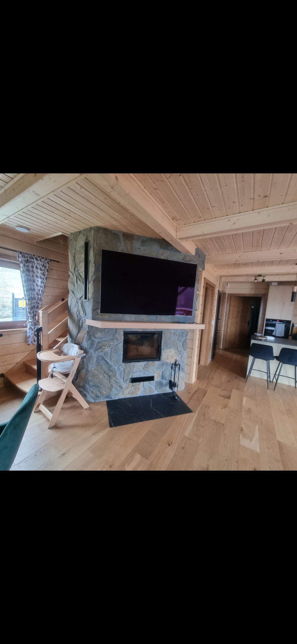 Drewniany salon z kamiennym kominkiem, na którym wisi telewizor. Widoczny drewniany sufit, podłoga i schody z dziecięcym krzesełkiem. Czarny marmurowy element pod kominkiem.