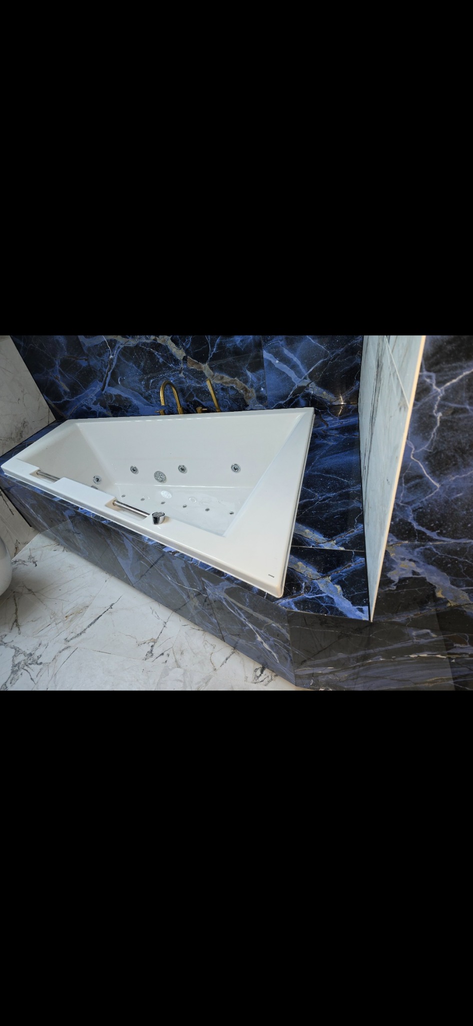 Nowoczesna, biała wanna z hydromasażem w łazience wykończonej płytkami imitującymi marmur w odcieniach bieli i granatu, z baterią w kolorze złotym.