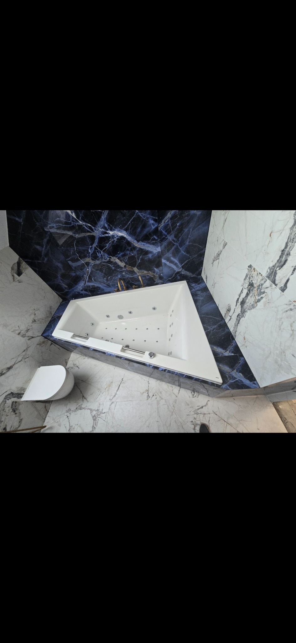 Nowoczesna łazienka z narożną wanną jacuzzi, wykończona marmurem w odcieniach bieli i granatu, z widoczną toaletą i złotą armaturą.