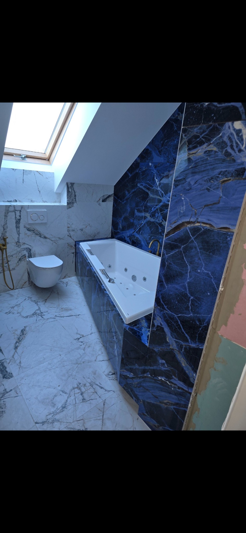 Biała wanna z hydromasażem obudowana ciemnoniebieskimi płytkami imitującymi marmur, z widoczną białą toaletą i oknem dachowym w wykończonej łazience na poddaszu.