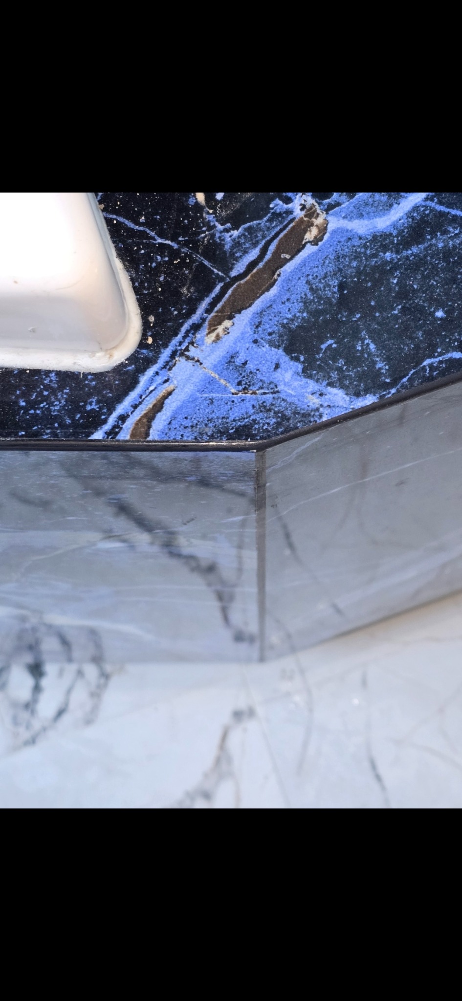 Fragment wykończonej łazienki z umywalką, blatem z czarnego marmuru z niebieskimi żyłami i białymi płytkami podłogowymi z szarymi wzorami.