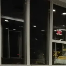 Aluminiowa drabina oparta o okno z naklejką ostrzegawczą 'Bezwzględny zakaz otwierania okien', widoczny niebieski pojemnik i okna budynku biurowego.