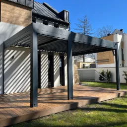 Nowoczesna pergola metalowa w kolorze antracytowym, z lamelami na dachu, przylegająca do domu z drewnianą elewacją, na drewnianym tarasie, w słoneczny dzień.