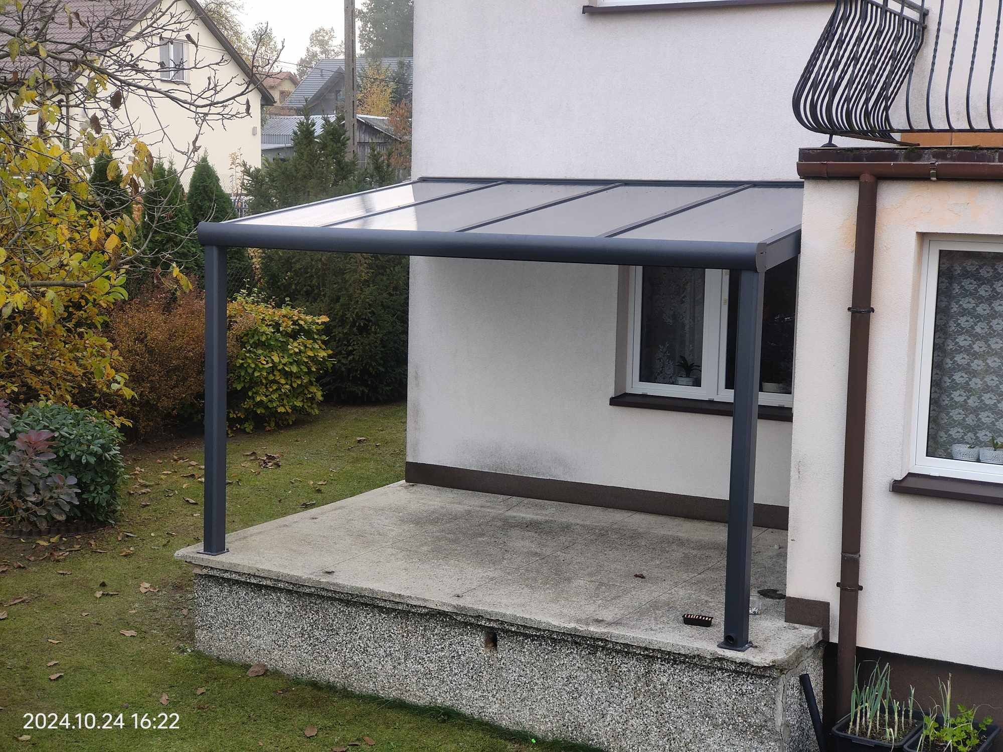 Zadaszony taras z metalową konstrukcją w kolorze antracytowym, przylegający do jasnego budynku z widocznym oknem i balkonem z kutą balustradą. Na pierwszym planie roślinność jesienna.