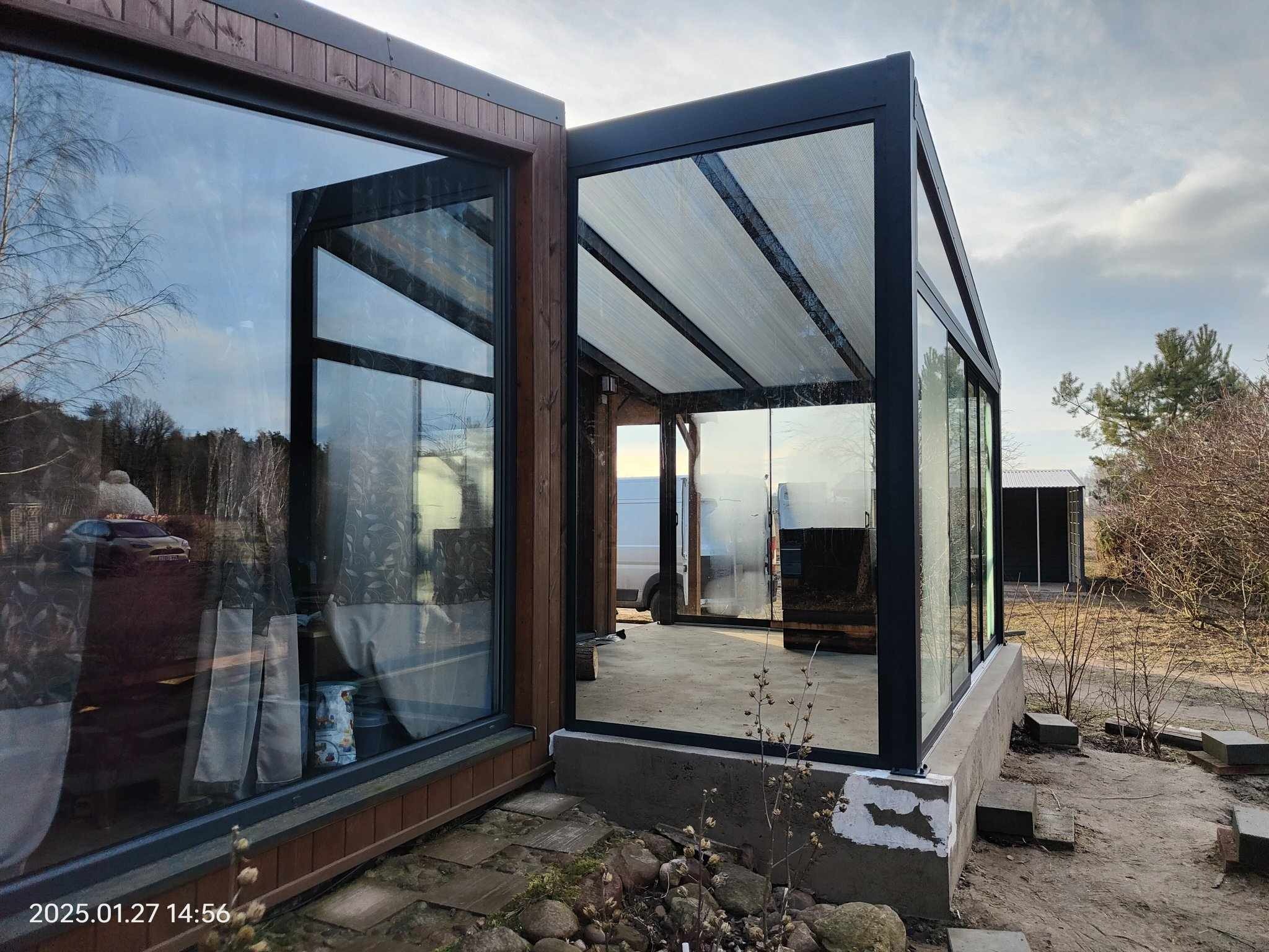 Metalowa konstrukcja z przeszklonymi ścianami i zadaszeniem na zewnątrz budynku, widok z boku, w tle drzewa i niebo.