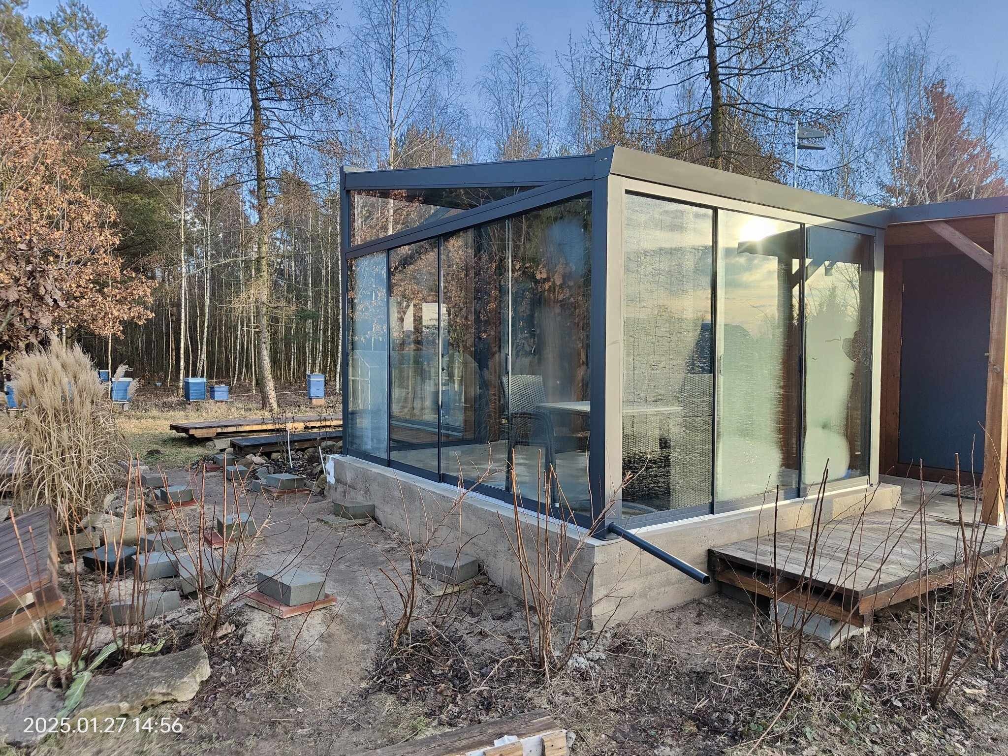 Nowoczesna przeszklona konstrukcja tarasowa z metalową ramą przylegająca do budynku, z widocznymi ulami w tle, w trakcie prac wykończeniowych terenu.