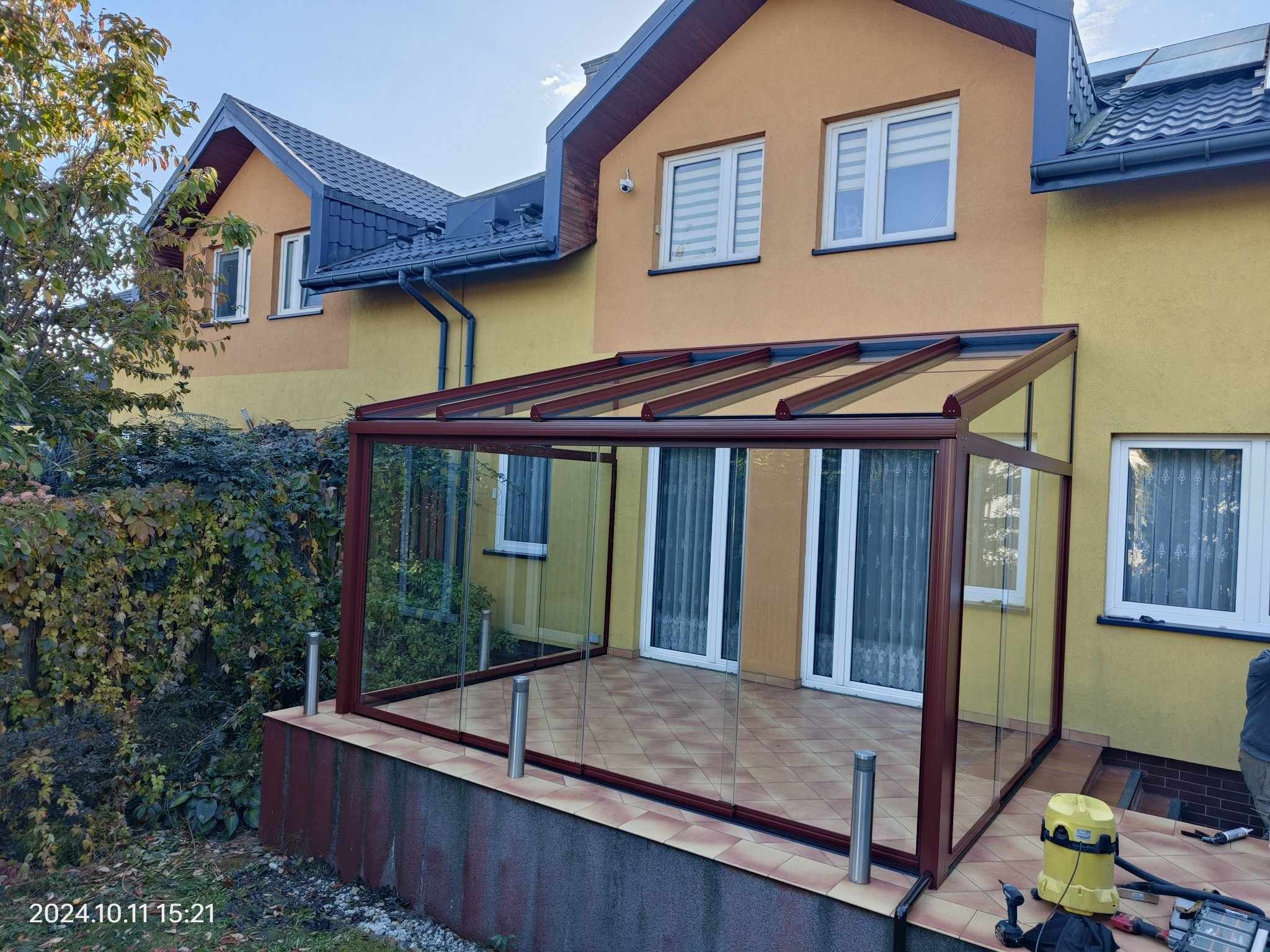 Zadaszona metalowa pergola z przezroczystymi ścianami, brązowa rama, zamontowana przy dwukondygnacyjnym domu z żółtą elewacją i szarą dachówką, widoczna część narzędzi i odkurzacza po prawej stronie.