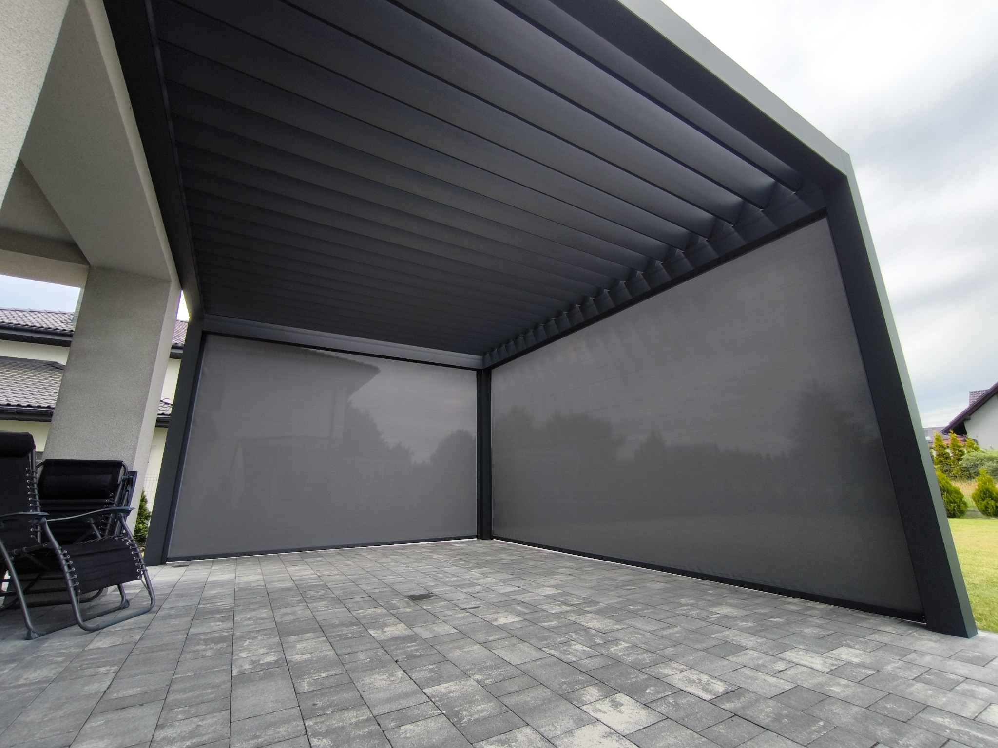 Nowoczesna, szara pergola z metalową konstrukcją i zwijanymi przesłonami przeciwsłonecznymi, ustawiona na brukowanym patio obok domu.