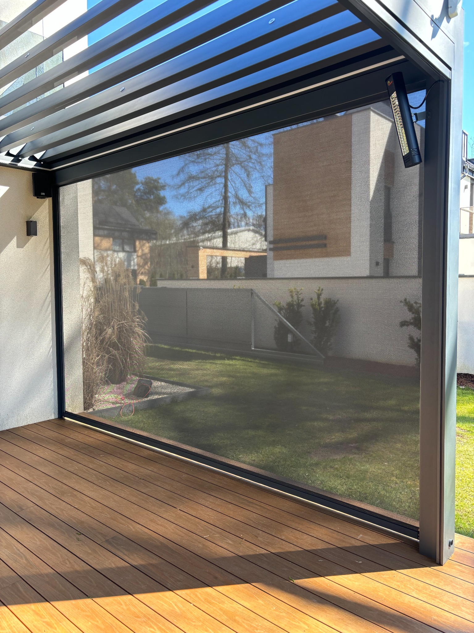 Nowoczesna, metalowa pergola z zasłoniętą roletą screen, widok na ogród z trawnikiem i krzewami, oświetlenie zewnętrzne zamontowane na konstrukcji, deski tarasowe na podłodze.