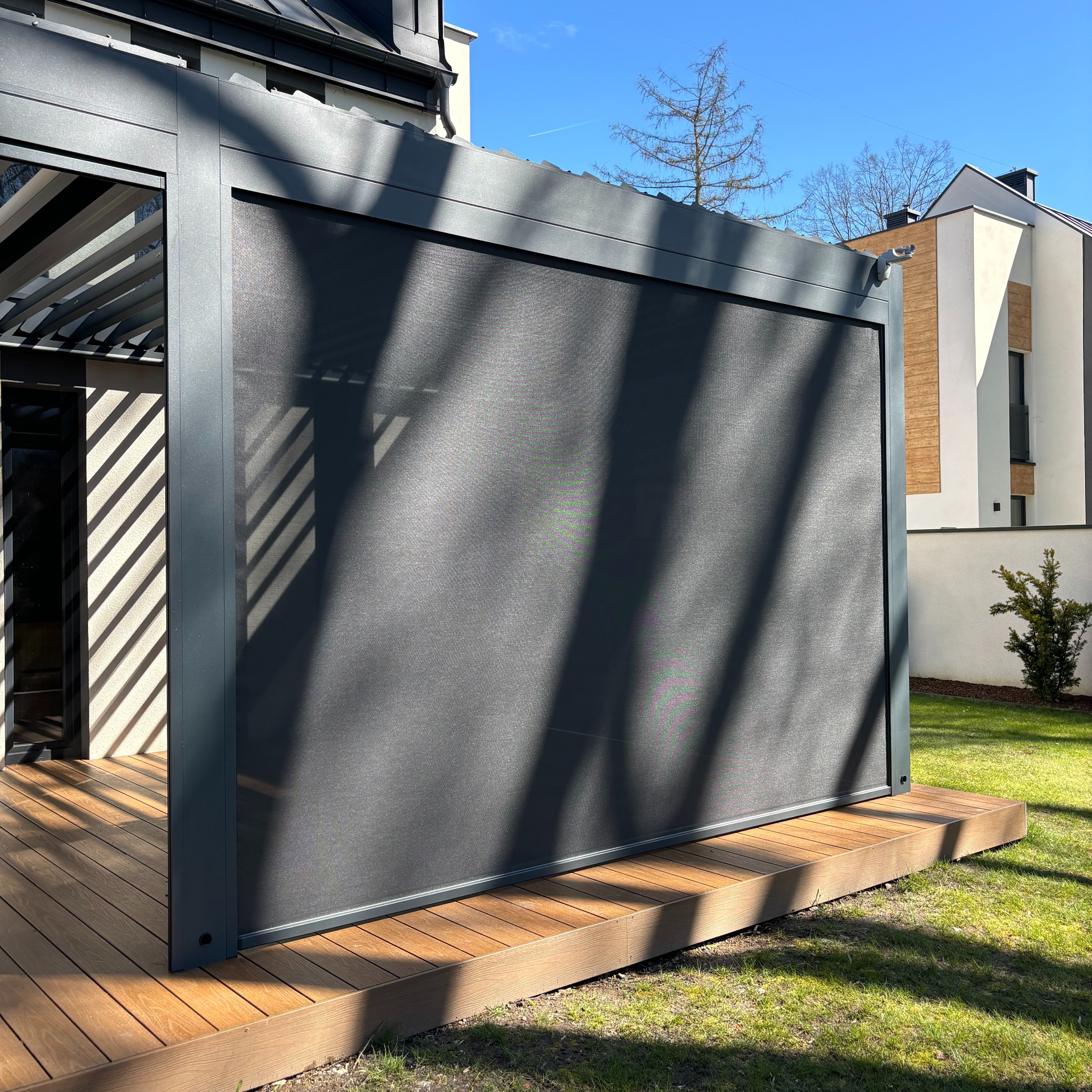 Nowoczesna pergola z metalową konstrukcją i roletą screenową zacieniającą taras z desek kompozytowych, w tle budynek mieszkalny i błękitne niebo.