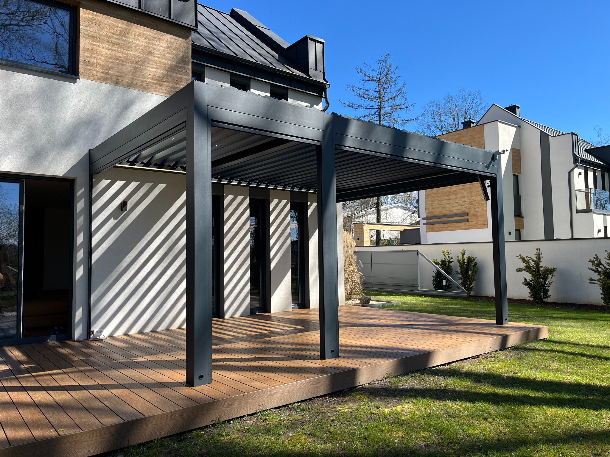Nowoczesna pergola metalowa w kolorze antracytowym, z lamelami na dachu, przylegająca do domu z drewnianą elewacją, na drewnianym tarasie, w słoneczny dzień.