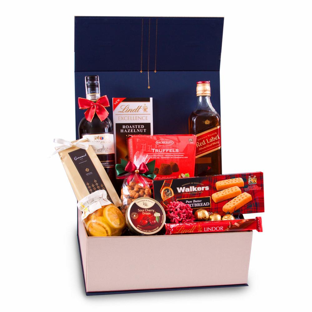 Otwarty, elegancki granatowo-różowy karton z selekcją wysokiej jakości produktów spożywczych, w tym butelki wina i whisky z czerwonymi kokardami, czekolady Lindt, trufle, ciastka Walkers i słoik...