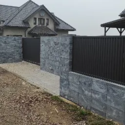 Nowoczesna posesja z przesuwną bramą wjazdową i ogrodzeniem panelowym na podmurówce z bloczków betonowych, widoczny znak 'Obiekt Monitorowany'.