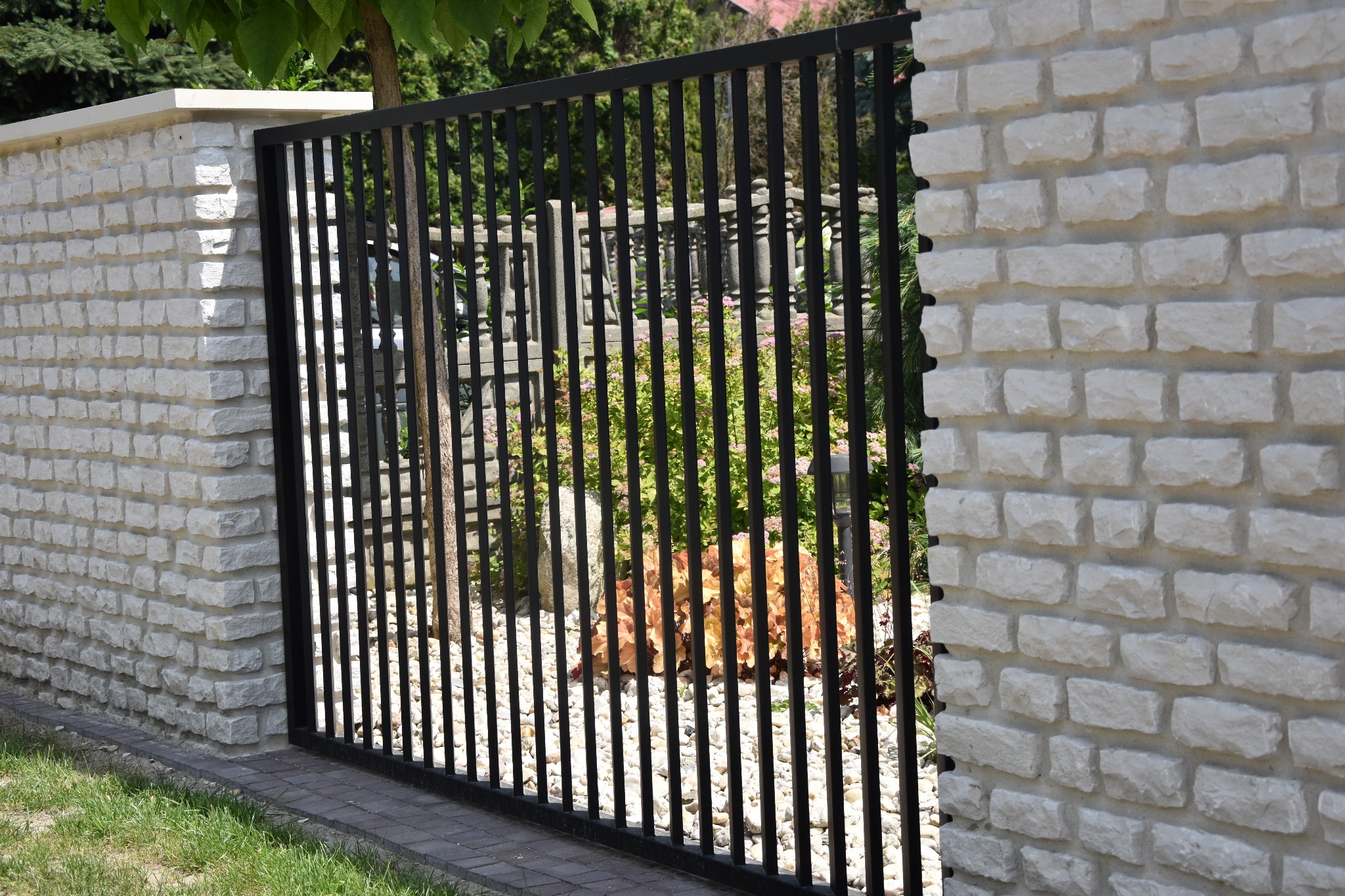 Nowoczesna, czarna brama wjazdowa z pionowych profili, osadzona w murze z białej cegły w Rudnikach, z widokiem na zadbany ogród.