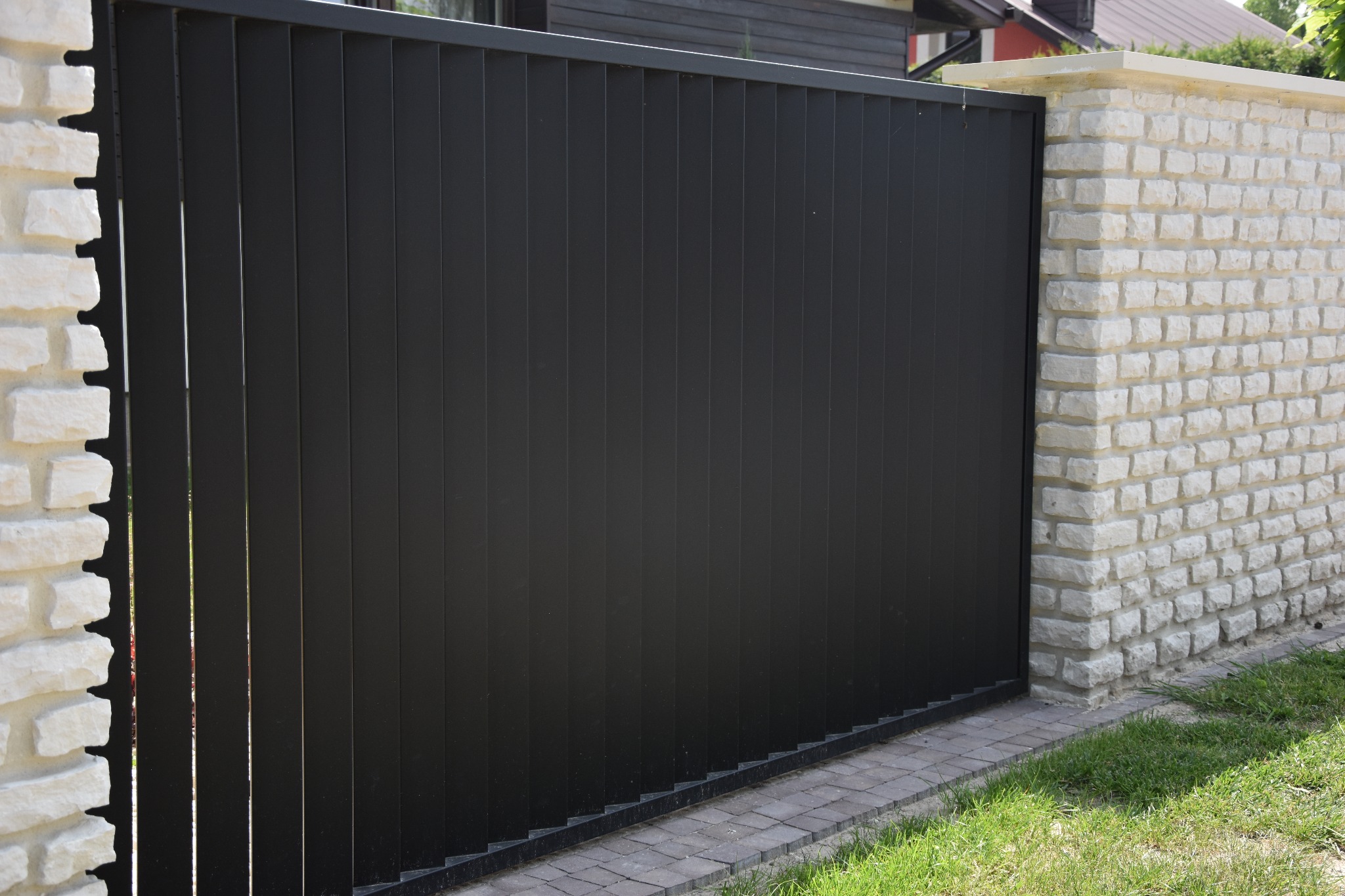 Nowoczesna brama przesuwna z czarnych, pionowych paneli, osadzona między słupkami z jasnego kamienia łupanego, z kostką brukową i trawą u podstawy.
