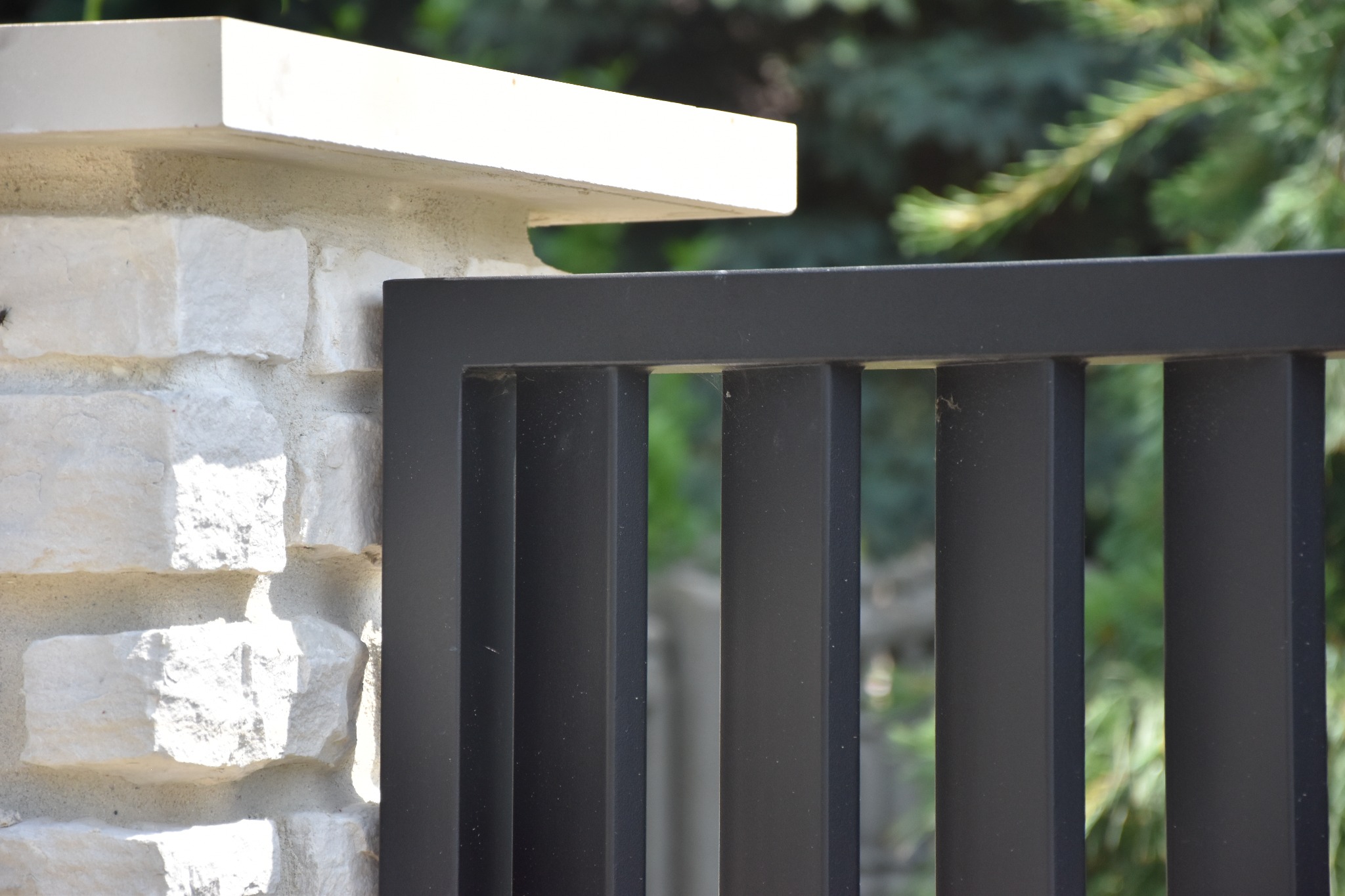 Fragment nowoczesnego ogrodzenia: czarne, pionowe metalowe panele przymocowane do słupka z jasnego kamienia z betonowym zwieńczeniem, z zielenią w tle.