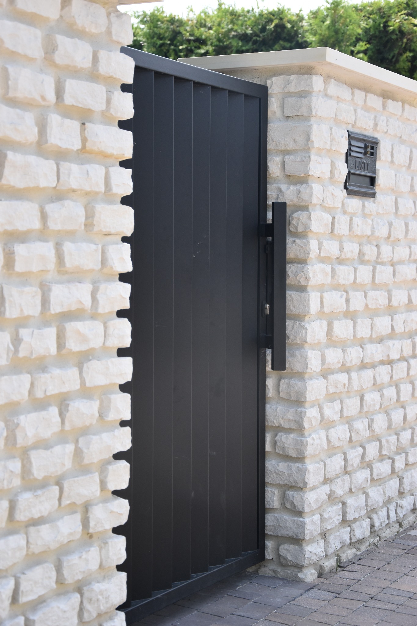 Nowoczesna, pionowa brama w kolorze antracytowym, zamontowana w murze z jasnego kamienia łupanego, z minimalistyczną czarną klamką i skrzynką na listy.