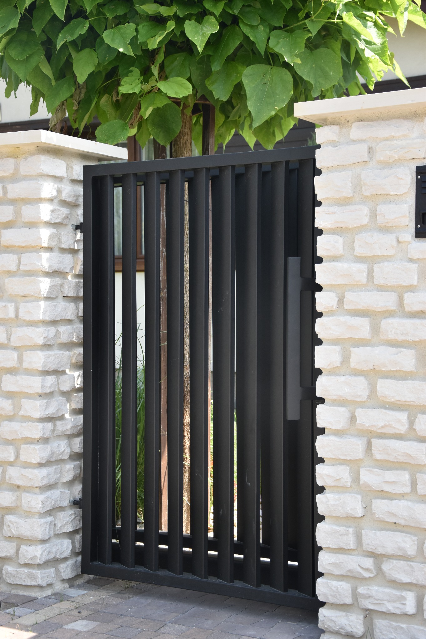 Nowoczesna brama wjazdowa z czarnych, pionowych profili, osadzona w białym murze z kamienia łupanego, z widocznym fragmentem drzewa w tle.