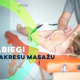 Terapeuta wykonuje zabieg na plecach pacjentki leżącej na pomarańczowym stole do zabiegów, widoczne dłonie terapeuty i fragment ucha pacjentki z kolczykiem.