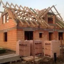 Budowa domu jednorodzinnego w trakcie wznoszenia ścian z bloczków ceramicznych, widoczna konstrukcja dachu i palety z materiałem budowlanym