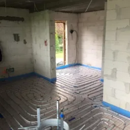 Rozłożona instalacja ogrzewania podłogowego w surowym pomieszczeniu, rury grzewcze ułożone w spiralny wzór na izolacji, widoczny rozwijak do rur i surowe ściany z betonu komórkowego.