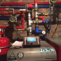 Kotłownia w Dobre Miasto z piecem na pellet KSR Beta Pellet 16, widoczne rury instalacji centralnego ogrzewania w kolorach czerwonym i niebieskim oraz ceglana ściana w tle.