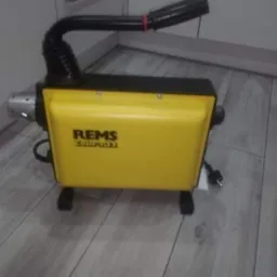 Żółta maszyna REMS Cobra 120 do czyszczenia rur, z czarnym wężem i zasilaniem, stojąca na panelach podłogowych.