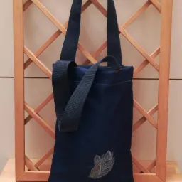 Granatowa torba na ramię z haftowanym motywem pióra pawia, ustawiona na drewnianym blacie przed ażurową kratownicą.