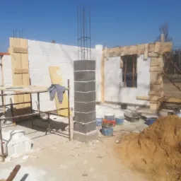 Wznoszenie ścian z bloczków betonu komórkowego na budowie domu, widoczne zbrojenie kolumny i drewniane szalunki okien, sterta piasku i narzędzia budowlane.