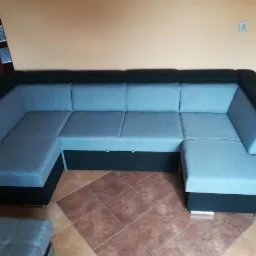 Rozłożysty narożnik tapicerowany szarą tkaniną z czarnymi akcentami, widoczne schowki pod siedziskami, brązowa podłoga z płytek.