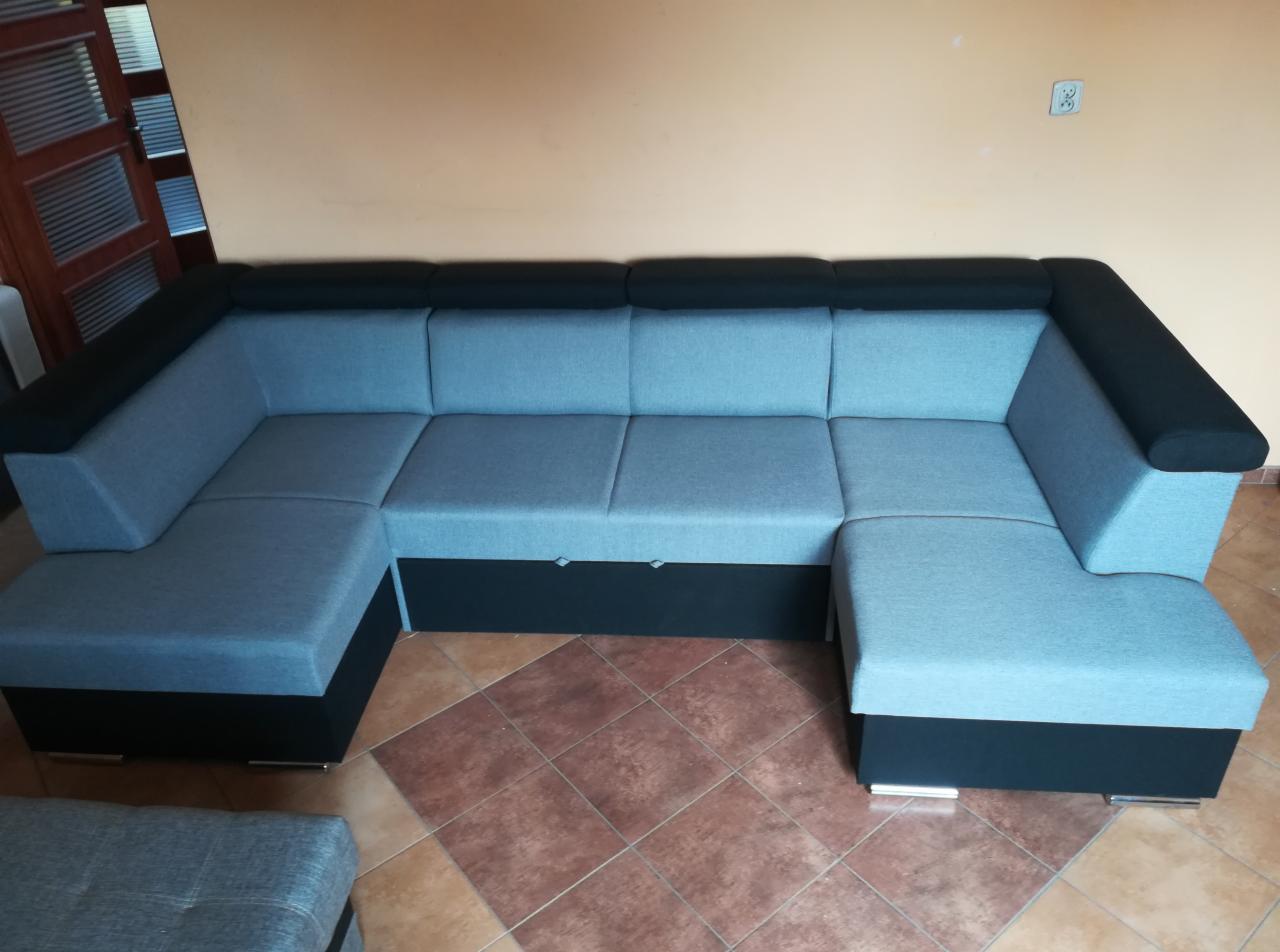 Szaro-czarny narożnik z funkcją spania i pojemnikiem na pościel, widoczny na tle pomarańczowej ściany i brązowej podłogi z płytek.