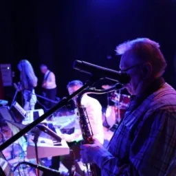 Saksofonista w koszuli w kratę gra solo na scenie podczas występu zespołu muzycznego, widoczni inni muzycy i instrumenty w tle, oświetlenie sceniczne.