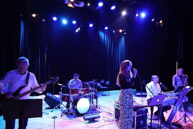 Zespół muzyczny na scenie: wokalistka w zebrę, basista w białej koszuli, perkusista, klawiszowiec z laptopem Apple i saksofonista, oświetlenie sceniczne w odcieniach niebieskiego.