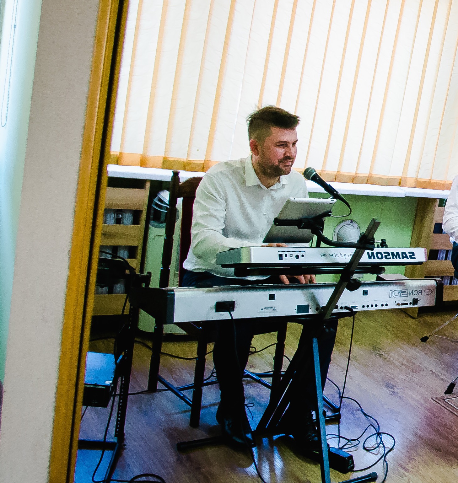 Mężczyzna w białej koszuli grający na keyboardzie Samson z tabletem i mikrofonem, widoczne kable i drewniane krzesło, ujęcie z boku.