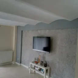 Pokój dzienny po remoncie: dekoracyjna ściana z efektem struktury, telewizor na ścianie, białe listwy przypodłogowe, grzejnik, sufit z dekoracyjnym wykończeniem, białe zasłony.