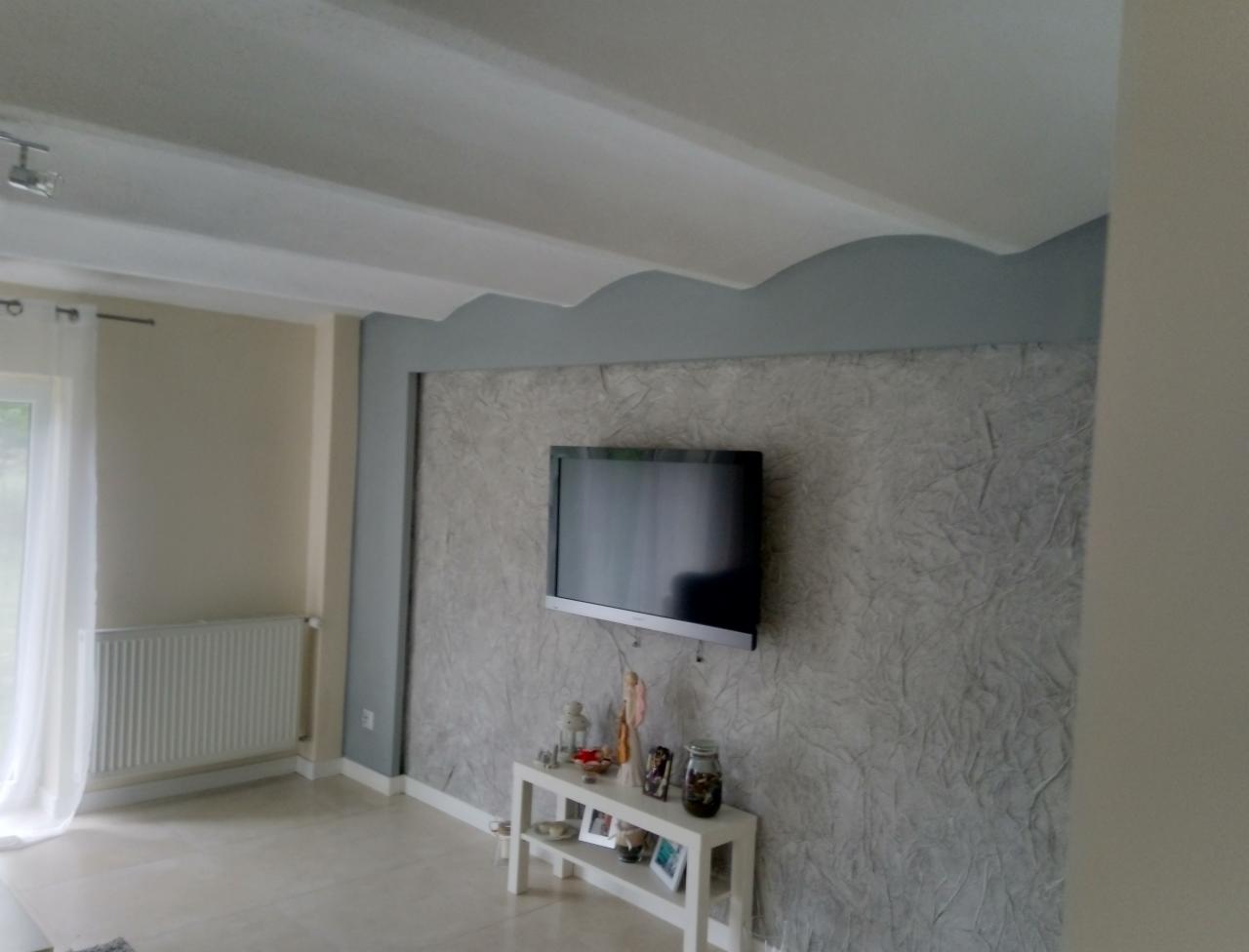 Pokój dzienny po remoncie: dekoracyjna ściana z efektem struktury, telewizor na ścianie, białe listwy przypodłogowe, grzejnik, sufit z dekoracyjnym wykończeniem, białe zasłony.