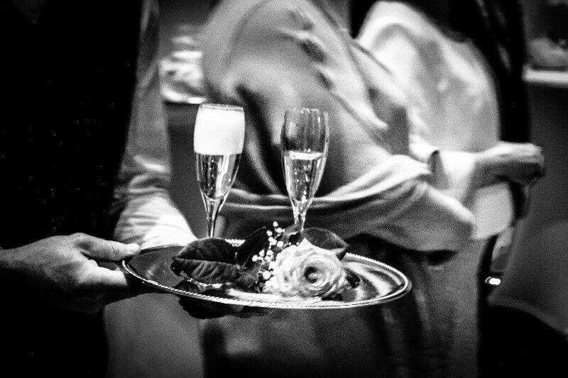 Czarno-białe zdjęcie kelnera niosącego srebrną tacę z dwoma kieliszkami szampana i dekoracją z róży oraz liści.