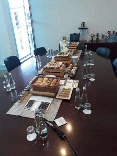 Długi stół konferencyjny z brązowego drewna, zastawiony przekąskami, napojami i sztućcami na jasnych serwetach, przygotowany na spotkanie biznesowe lub event.