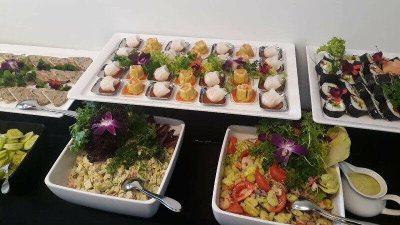 Bufet z różnorodnymi przekąskami: sushi, sałatki, terrina i pierożki dim sum, udekorowane kwiatami i zielenią, na białych naczyniach.