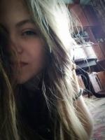 Portret młodej kobiety z długimi blond włosami, leżącej na wzorzystym dywanie, z głową opartą na ręce, w nieostrym tle widać fragment drewnianego mebla.