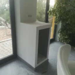 Narożny element zabudowy z wnęką, prawdopodobnie wykonany z płyt gipsowo-kartonowych, z widocznymi śladami po gniazdku elektrycznym, na tle okna z widokiem na zewnątrz i rośliny doniczkowej.