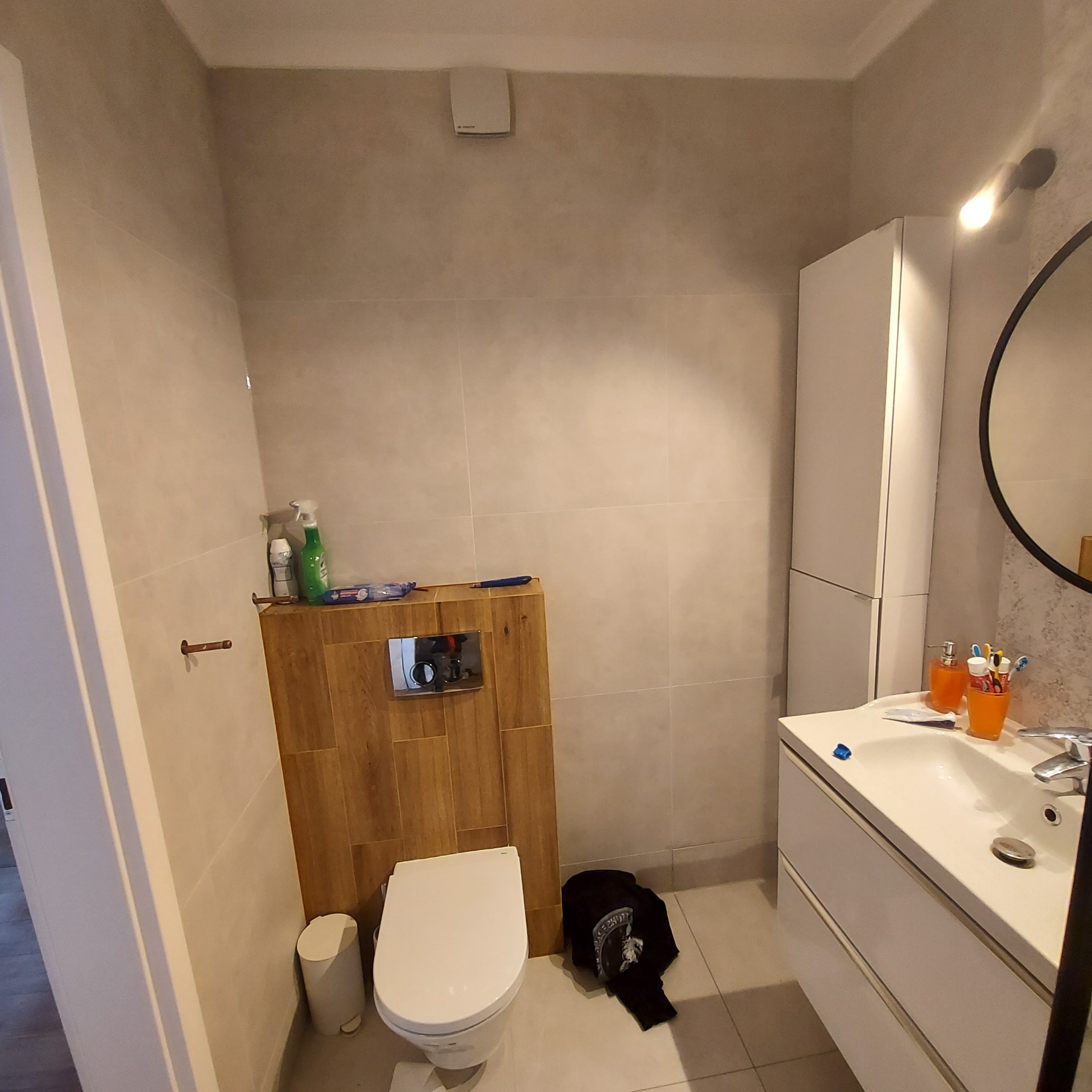 Wykończona łazienka z toaletą podwieszaną obudowaną drewnopodobnymi płytkami, umywalką z szafką w kolorze białym i wiszącą szafką. Na ścianie widoczny wentylator.