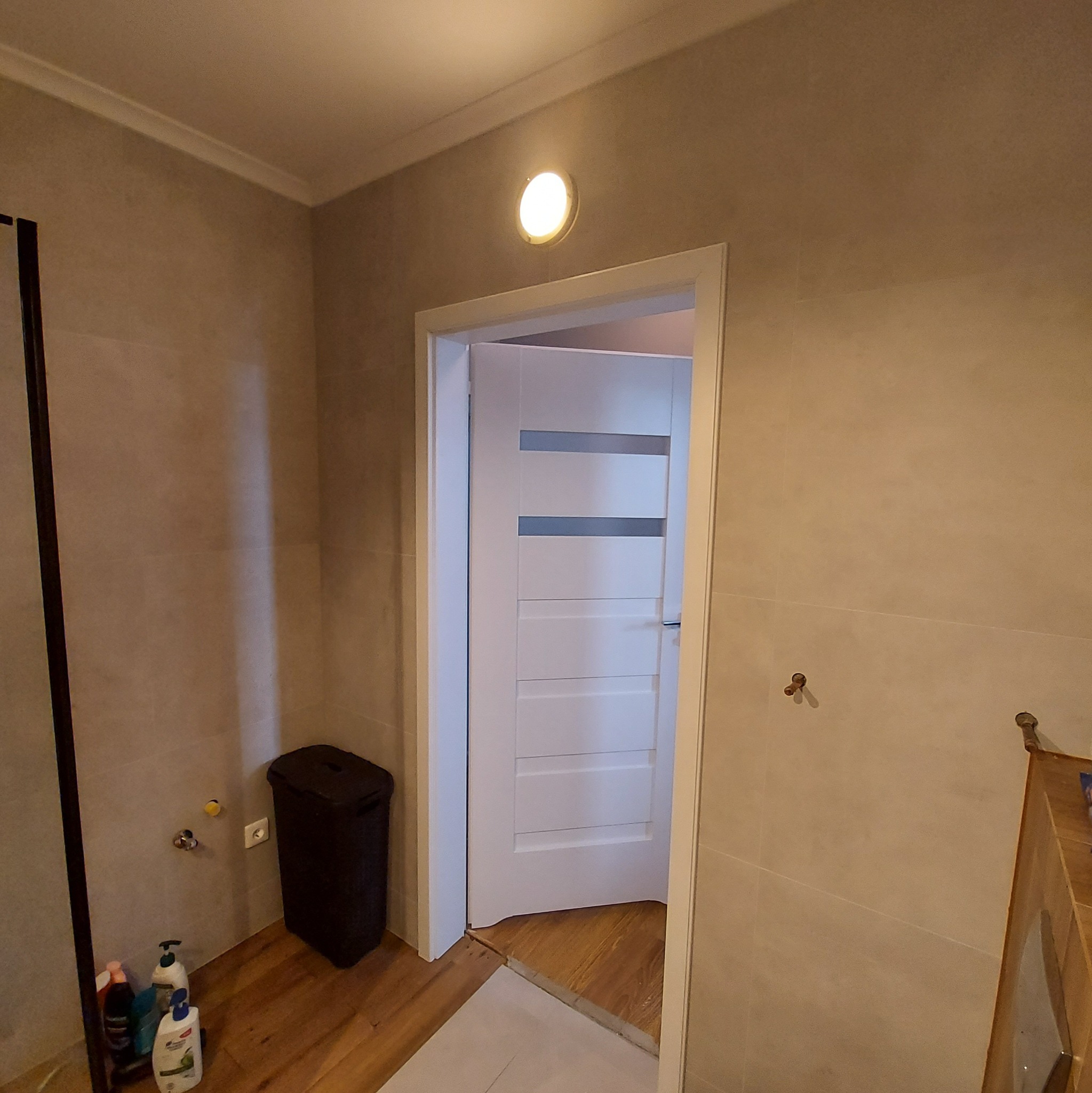 Wykończona łazienka z beżowymi płytkami, widoczna biała, nowoczesna drzwi wewnętrzne z poziomymi przeszkleniami, czarny kosz na pranie, fragment kabiny prysznicowej i drewniana podłoga.