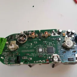 Zdjęcie od spodu deski rozdzielczej samochodu z widocznymi elementami elektronicznymi, w tym zielonymi i niebieskimi złączami, oraz centralnym procesorem, leżącej na białym stole.