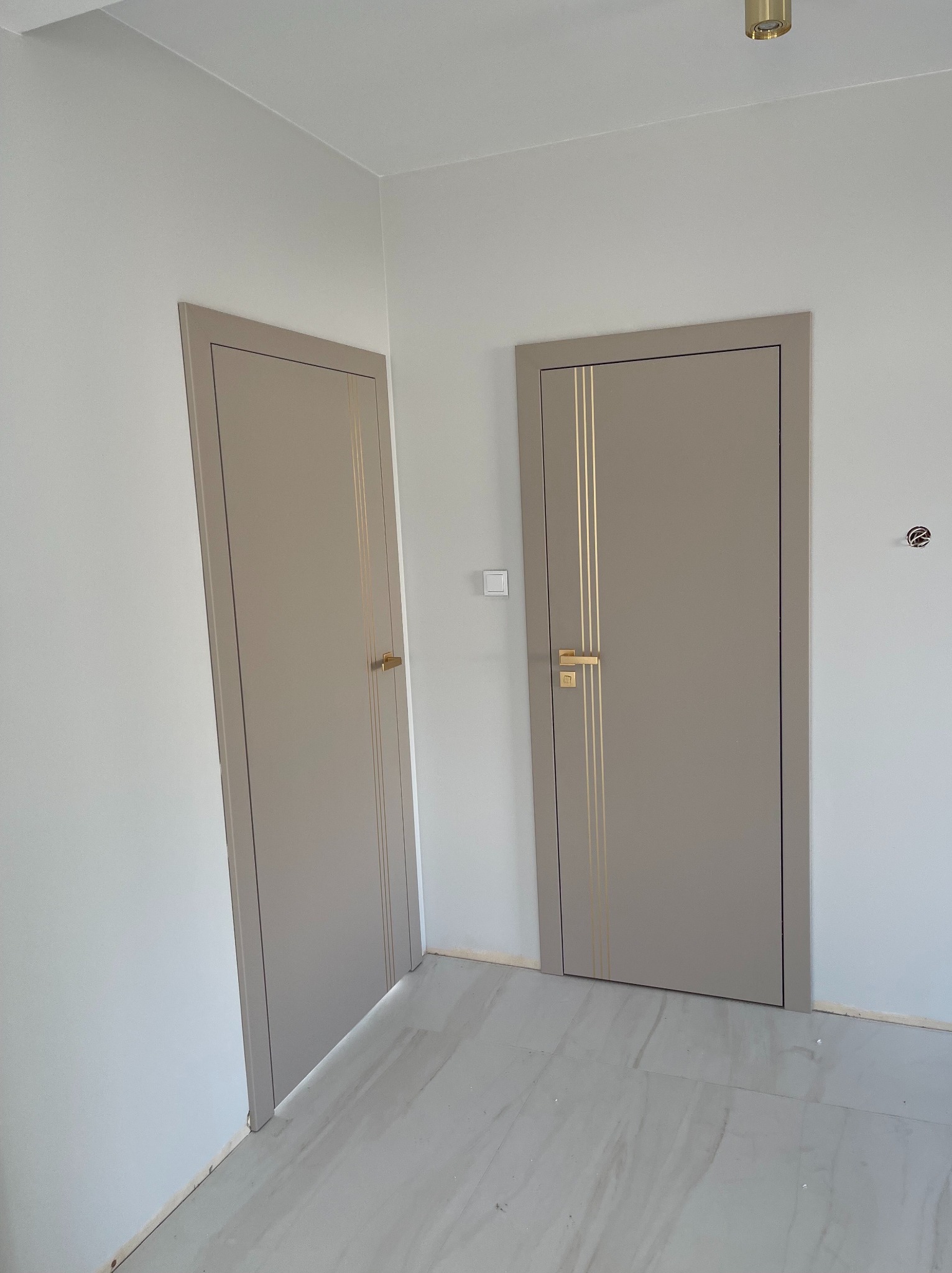 Dwa beżowe drzwi wewnętrzne z złotymi paskami i złotymi klamkami w nowym, jasnym wnętrzu. Jeden z nich jest lekko otwarty, ukazując fragment drugiego pomieszczenia.