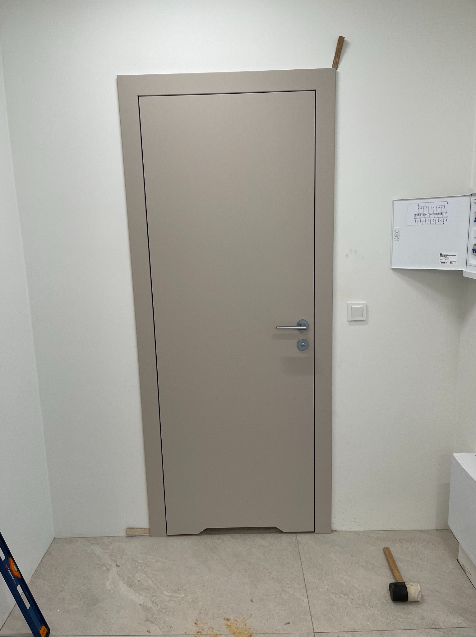 Zamontowane, beżowe drzwi wewnętrzne z srebrną klamką, widoczne narzędzia montażowe i fragment skrzynki elektrycznej na białej ścianie.