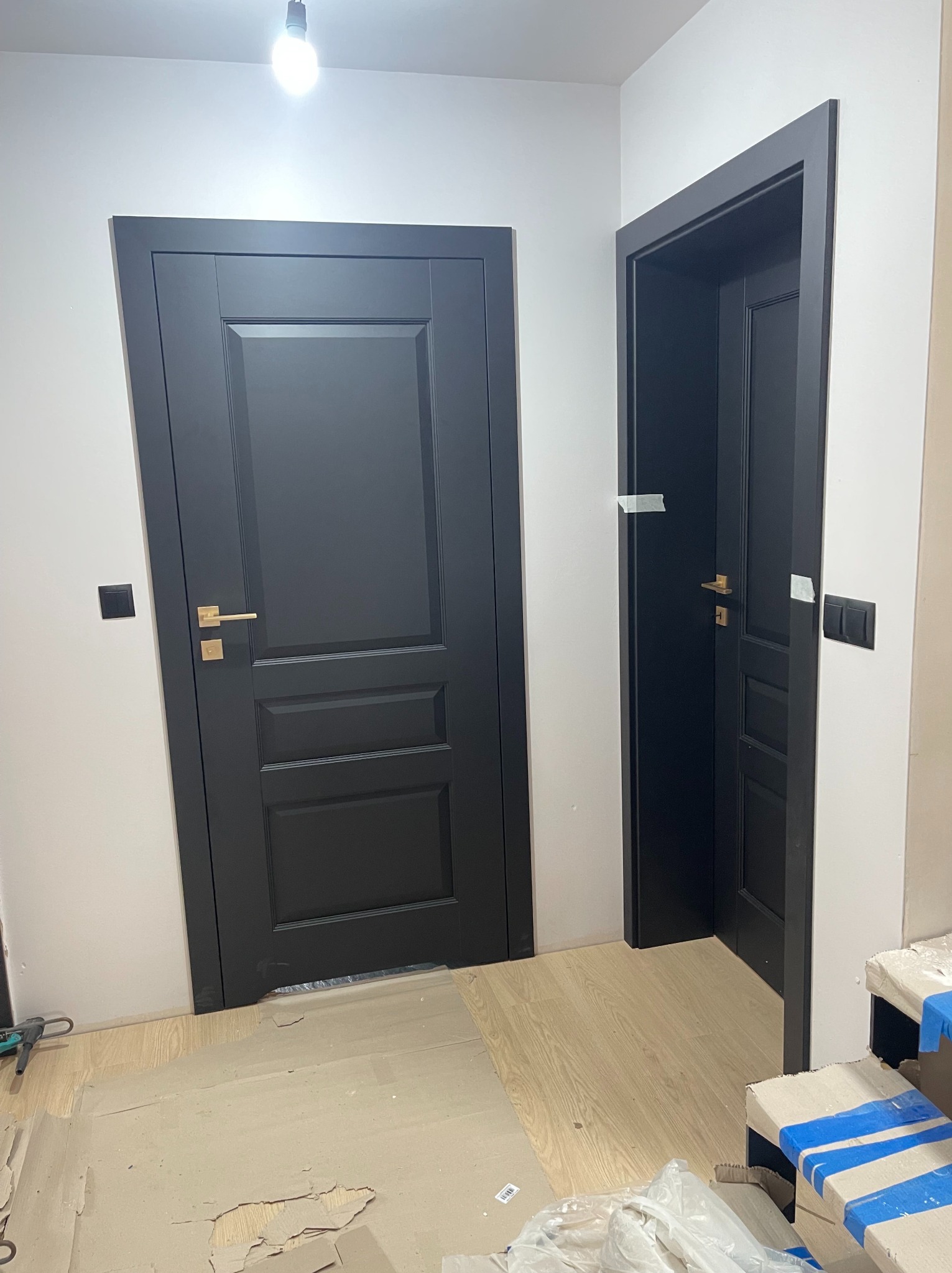Dwa czarne, klasyczne drzwi z złotymi klamkami, jeden otwarty, drugi zamknięty. Widoczne zabezpieczenia taśmą malarską. Podłoga zabezpieczona kartonem.