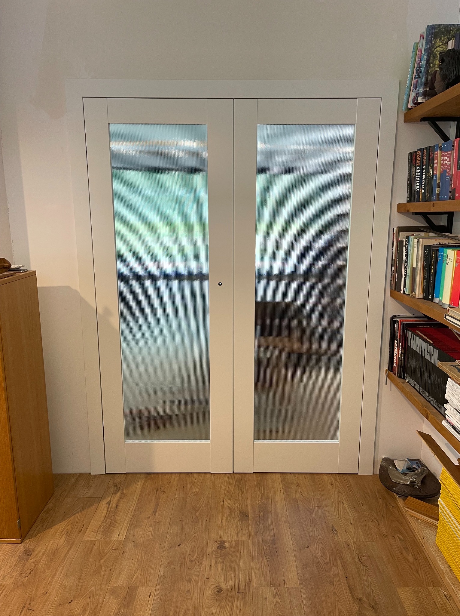 Białe, dwuskrzydłowe drzwi z ryflowanym szkłem, zamontowane w białej futrynie. Obok drewniany regał z książkami i drewniana podłoga.